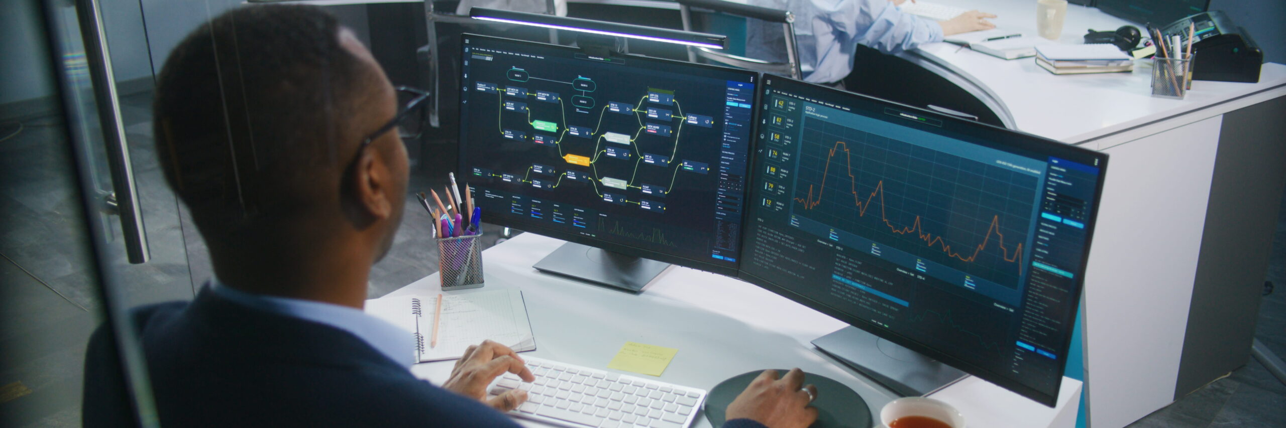 A data scientist in a suit works at a desk with three computer monitors displaying various data graphs, in a modern office setting.