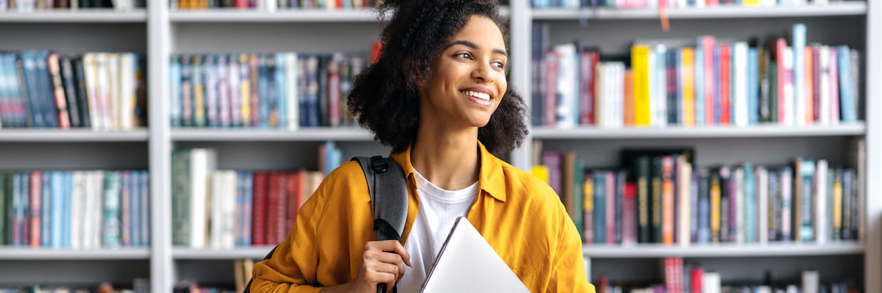 A young student with curly hair and a bright smile carrying a backpack and a folder, standing in a library with shelves full of books, ready to get started on her dream career.