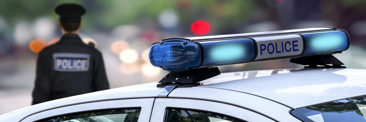 A police officer stands in front of a patrol car, focusing on the scene ahead. He is early in his police officer career, evident from the attentive way he surveys the area. The blue police lights are visible on top of the white vehicle, with blurred city lights in the background.