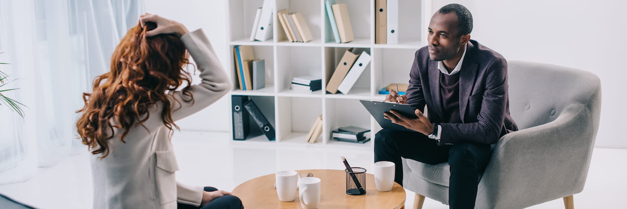 A psychiatrist listens attentively to a female client in distress during a counseling session in a modern office, with coffee cups on the table between them.
