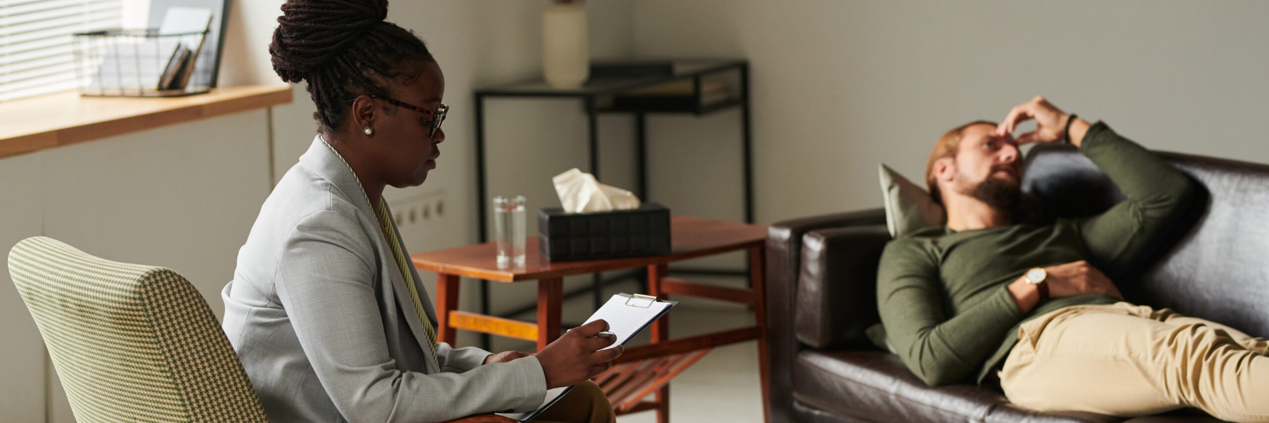 A psychologist, a black woman, takes notes while listening to a male patient lying thoughtfully on a couch in her office, evoking a counseling session.
