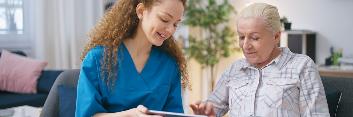 A young social worker in a blue uniform shows a tablet to an elderly woman seated beside her in a home setting, both smiling as they engage with the content.