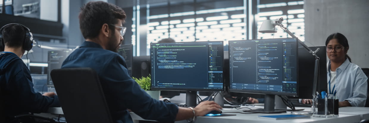 A modern office scene featuring three software developers at workstations with multiple monitors displaying code, focusing intently in a well-lit, high-tech environment.