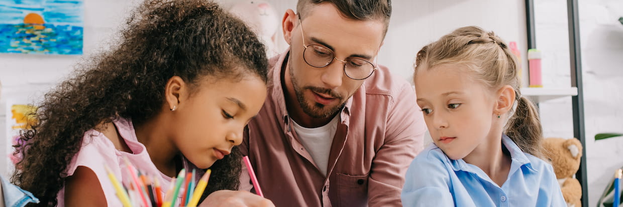 A teacher assists two young girls with their artwork in a colorful classroom, focusing on how to use colored pencils properly as part of a step-by-step guide.