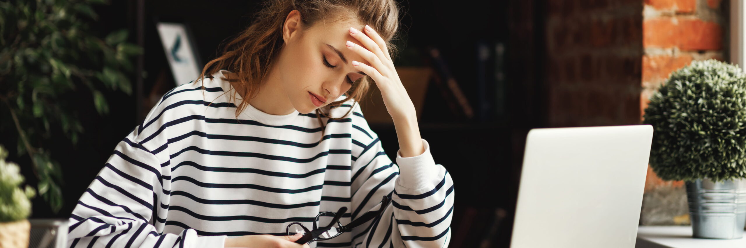 A woman in a striped top appears stressed or tired, holding her glasses and pressing her forehead while sitting at a table with a laptop in a cozy, brick-walled room. She could be reviewing the "Mental Health Handbook" for college students as she attempts to manage her workload and stress.