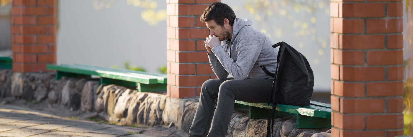 A student sits on a green park bench, looking at his phone with a serious expression, wearing a gray hoodie and black pants, with a backpack next to him.