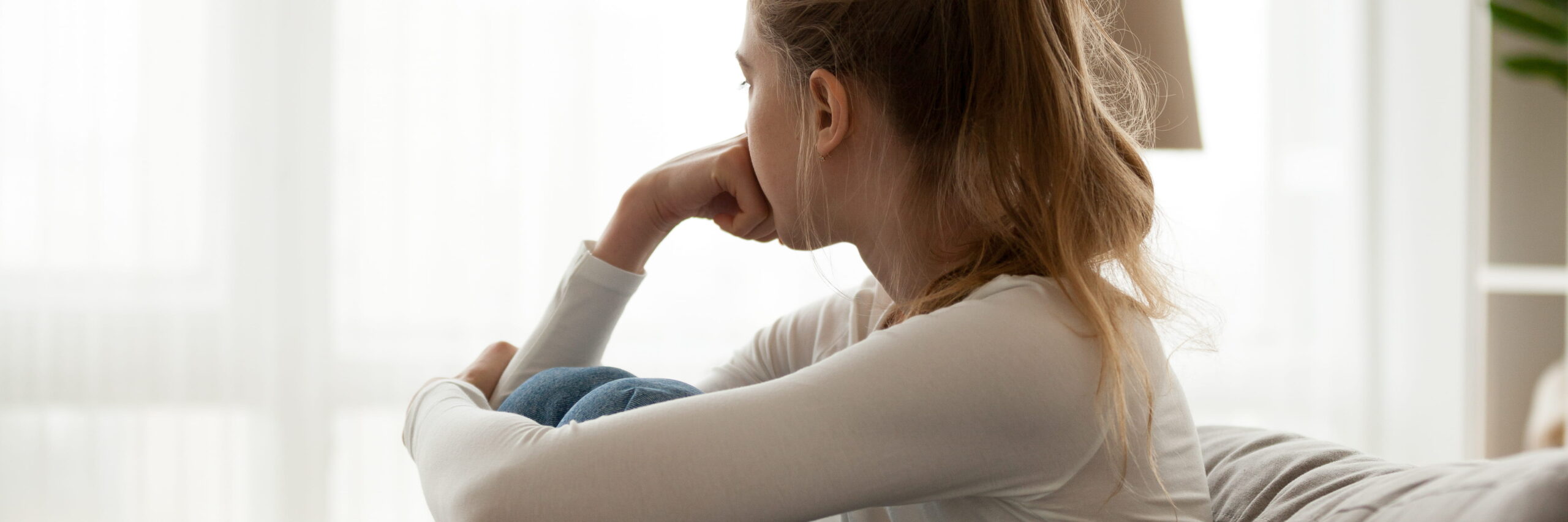 A woman sits pensively on a sofa, gazing out of a window with her chin resting on her hand, holding a blue fabric-draped arm, illuminated by soft natural light from behind. She reflects deeply on her college grief.

