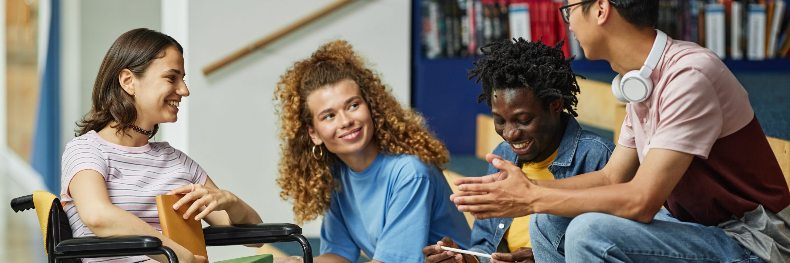 Three young students, one using a wheelchair, are engaging in a lively discussion about accessing education in a library setting, smiling and sharing thoughts.