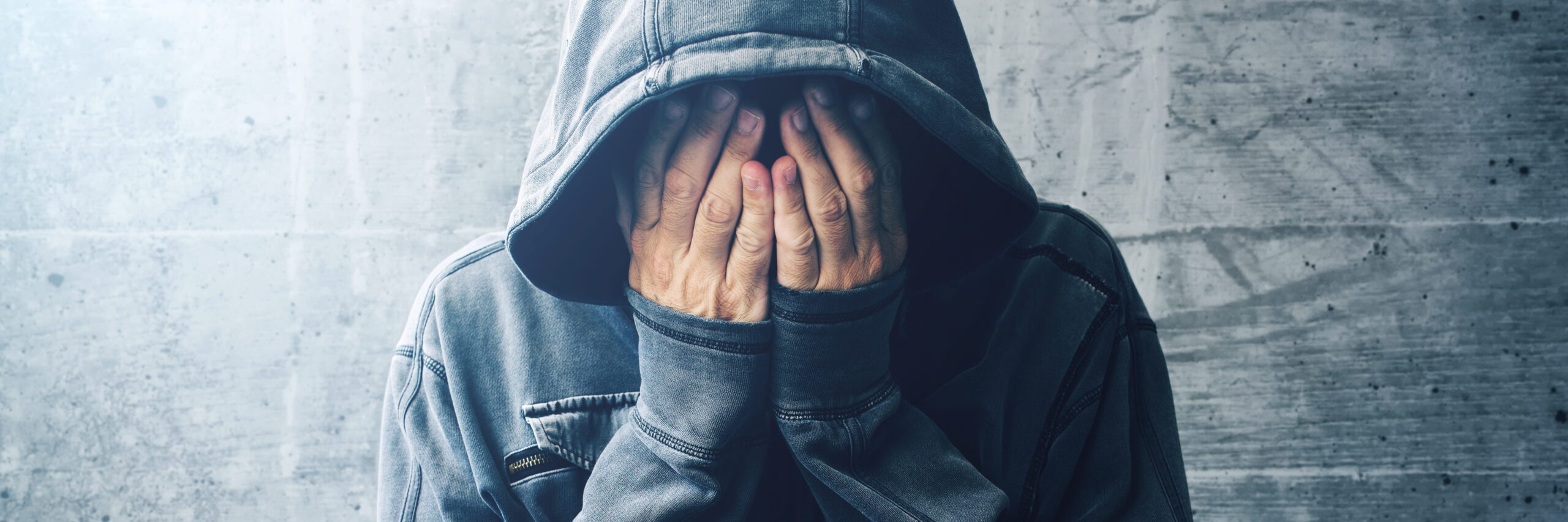 A college student in a gray hoodie covering their face with their hands against a textured concrete background, conveying a mood of sadness or contemplation.