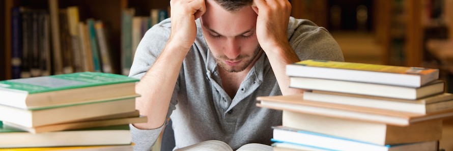 A focused man reading intently at a library table, surrounded by stacks of books, with shelves filled with more books in the background.